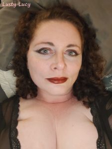 Lusty Lucy is wearing a dark top, showing lots of cleavage. She has a slight smile on her dark painted lips and dark eyeliner on!