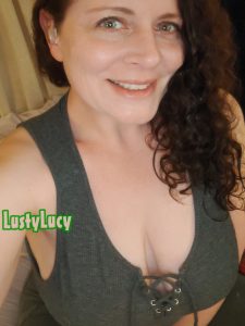 Cam model LustyLucy of LustyLucyPlays wearing a grey tank top, showing off her deep cleavage from her big boobs. She has her long curly hair swept to one side and big friendly smile on her face.